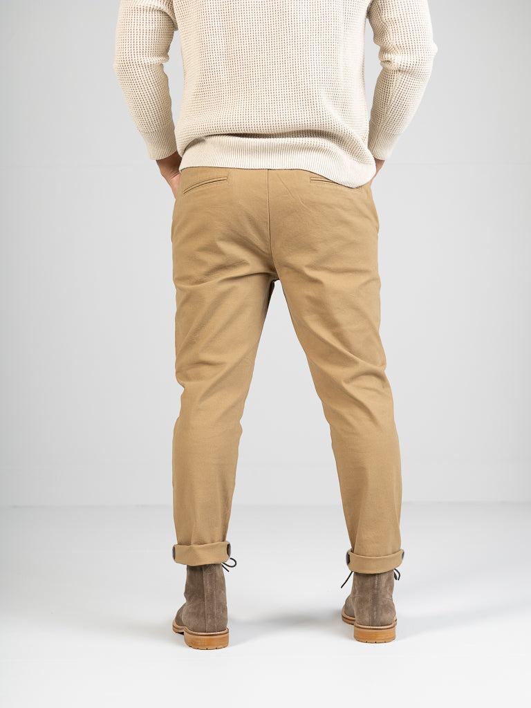Rear view of a person wearing a cream sweater, ID Menswears Kent Drawstring Canvas Pant in tan, and brown lace-up boots, standing against a plain light gray background.