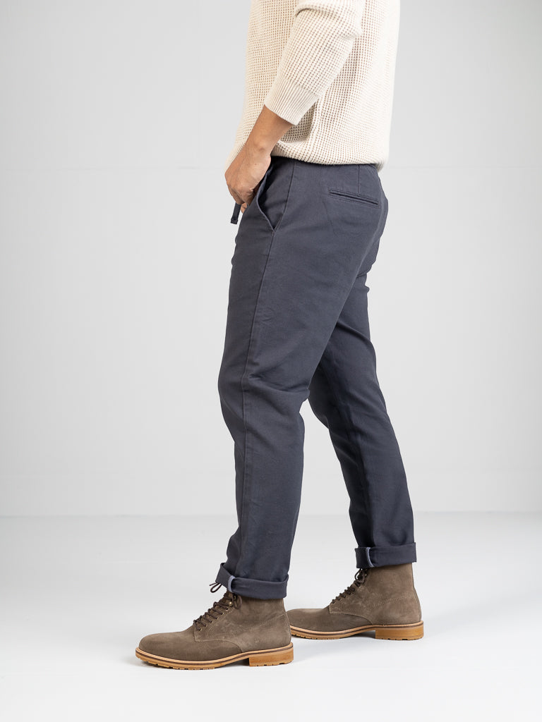 A person in a beige sweater and slim-fit ID Menswears Kent Drawstring Canvas Pant in dark gray stands in profile. They wear brown lace-up boots on a white background. The cotton canvas pants with cuffed hem seamlessly complete the casual look.
