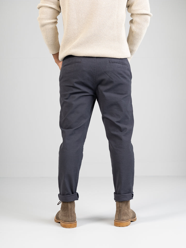 A rear view features someone in a cream knitted sweater, ID Menswears Kent drawstring canvas pants in gray, and brown suede ankle boots against a white background. Hands are casually tucked into pockets, completing the effortlessly chic look.