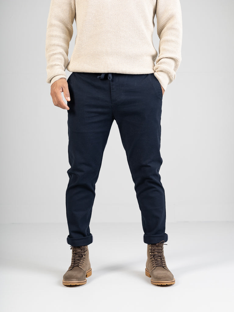 A person is seen against a plain white background wearing a cream-colored knit sweater, slim-fit navy blue Kent Drawstring Canvas Pants by ID Menswear with slightly rolled ankles, and light brown lace-up boots. The head is not visible.