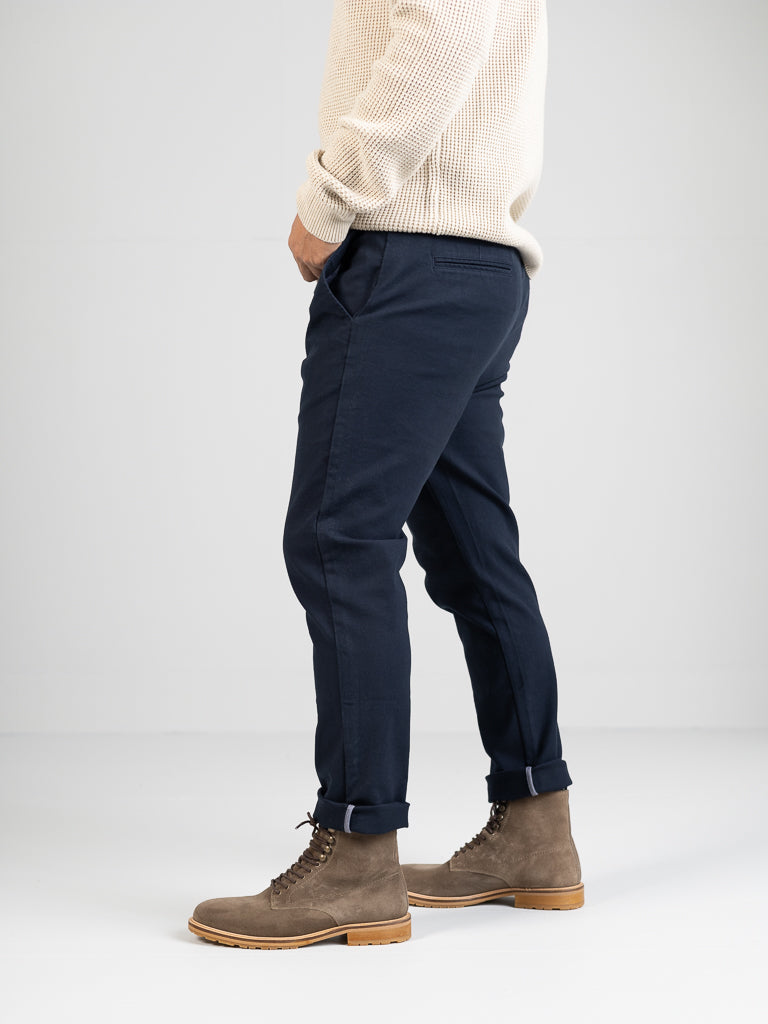 A person stands elegantly in a minimalistic, light-colored space, wearing the Kent Drawstring Canvas Pant by ID Menswear paired with a beige sweater and brown lace-up boots. Only their lower half is visible.