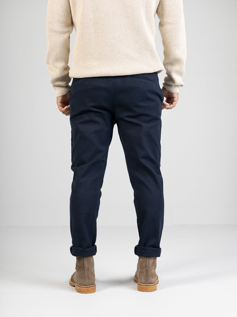 A person stands facing away in an ID Menswear cream sweater and dark blue Kent Drawstring Canvas Pant with a slim fit and mid-rise cuff design. Theyre also wearing light brown suede ankle boots, all against a plain white background.