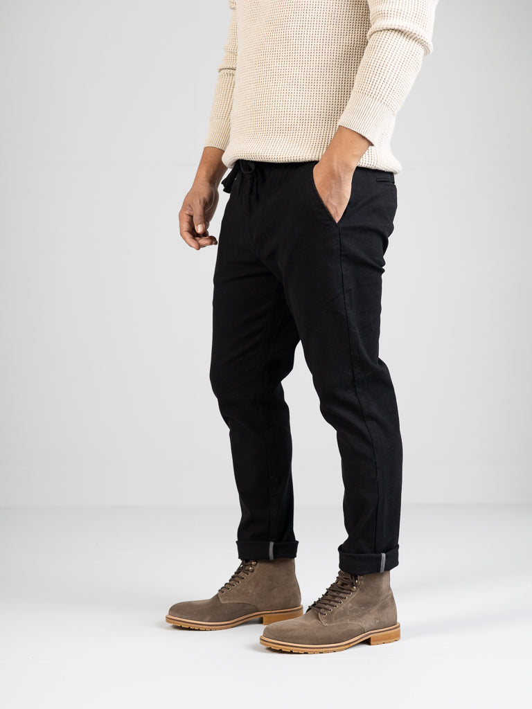 A person wearing the Kent Drawstring Canvas Pant from ID Menswear in black stands against a plain white background. They also wear a beige sweater and brown lace-up boots, with their hands tucked into the pants pockets. Their face is not visible.
