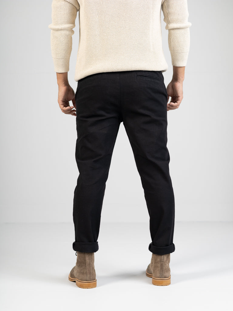 A person stands against a plain background, highlighting ID Menswears cream sweater with the Kent Drawstring Canvas Pant and brown suede boots, capturing the sleek silhouette of their style from behind.