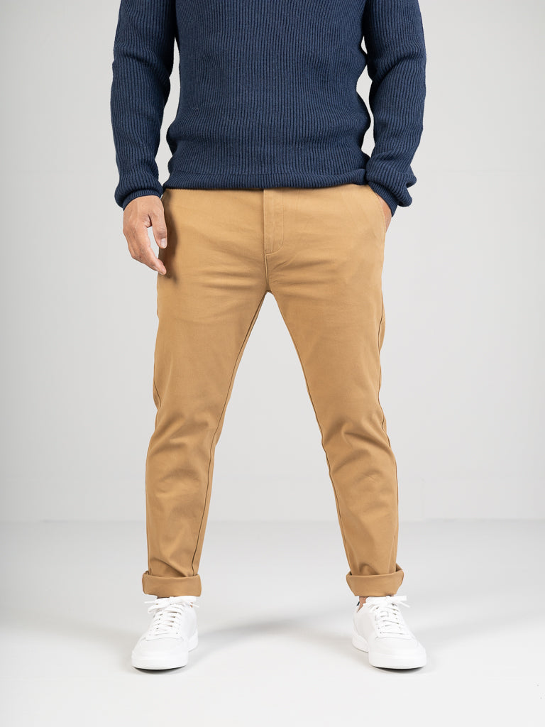 A person in a dark blue sweater, ID Menswears light brown Banker Twill Stretch Chino, and white sneakers stands against a plain light background with hands in pockets facing forward.