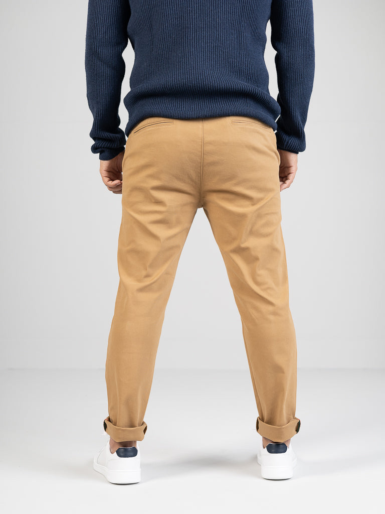 A person wearing a navy sweater, ID Menswears Banker Twill Stretch Chino, and white sneakers with dark accents stands facing away. The mid-rise chinos are regular slim-fit and slightly cuffed, set against a plain light gray background.