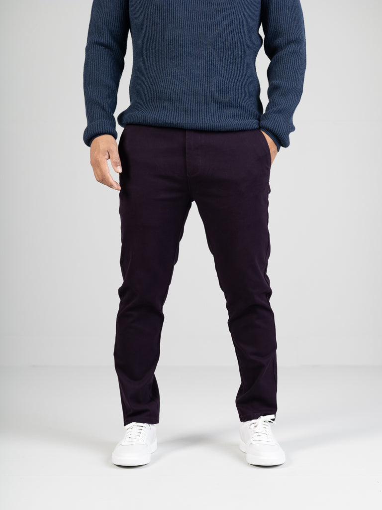 A person stands against a plain backdrop, sporting ID Menswears dark purple Banker Twill Stretch Chino with hands casually tucked into the pockets. Theyre also wearing a blue sweater and white sneakers.