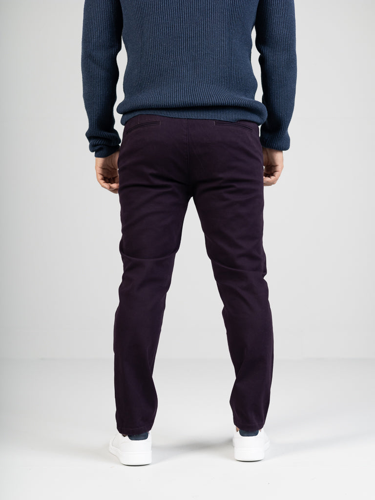 Rear view of someone wearing a blue sweater paired with ID Menswears Banker Twill Stretch Chino, regular slim-fit and mid-rise in dark purple, standing against a light gray background. White sneakers complete the look.