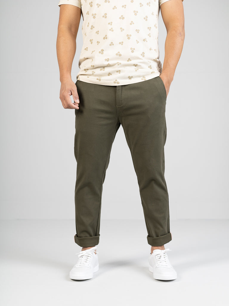 A person stands against a plain background, wearing a light patterned t-shirt, olive green regular slim-fit mid-rise Banker Twill Stretch Chino from ID Menswear, and white sneakers. Their face is hidden while hands are tucked in the pocket of their chinos.