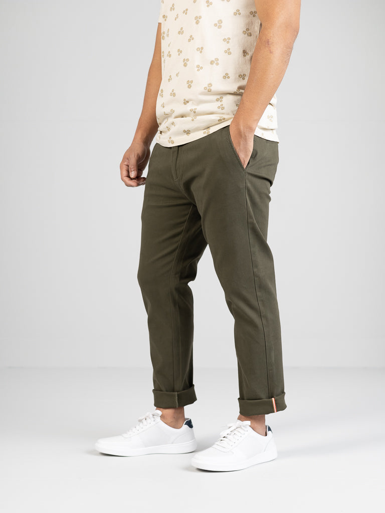 The image showcases someone in ID Menswears Banker Twill Stretch Chino, a mid-rise olive green pant with rolled cuffs, paired with a cream-colored floral pattern shirt and white sneakers, focusing on the lower half against a plain background.