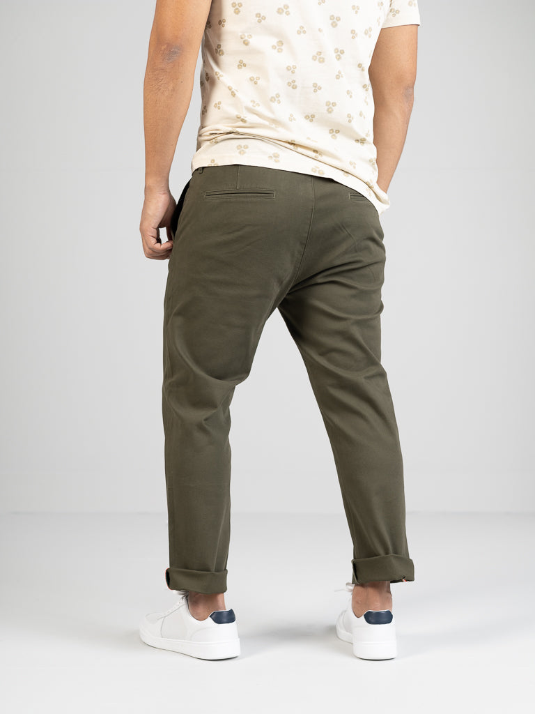 A person stands with their back to the camera, wearing an ID Menswear beige shirt with subtle patterns and dark green Banker Twill Stretch Chino pants. Their white sneakers contrast against a plain, light-colored backdrop.