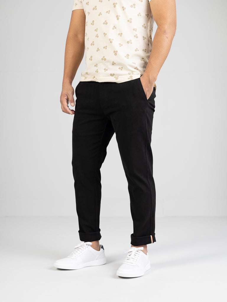 A person in a floral light-colored shirt, ID Menswears Banker Twill Stretch Chino pants, and white sneakers stands with hands in pockets against a neutral background. The outfit looks casual and stylish.