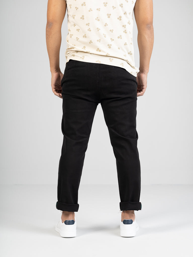 Rear view of a person in ID Menswears Banker Twill Stretch Chino, slim fit and black, paired with white sneakers and rolled cuffs. The ensemble is topped with a light-patterned t-shirt against a plain light gray background.