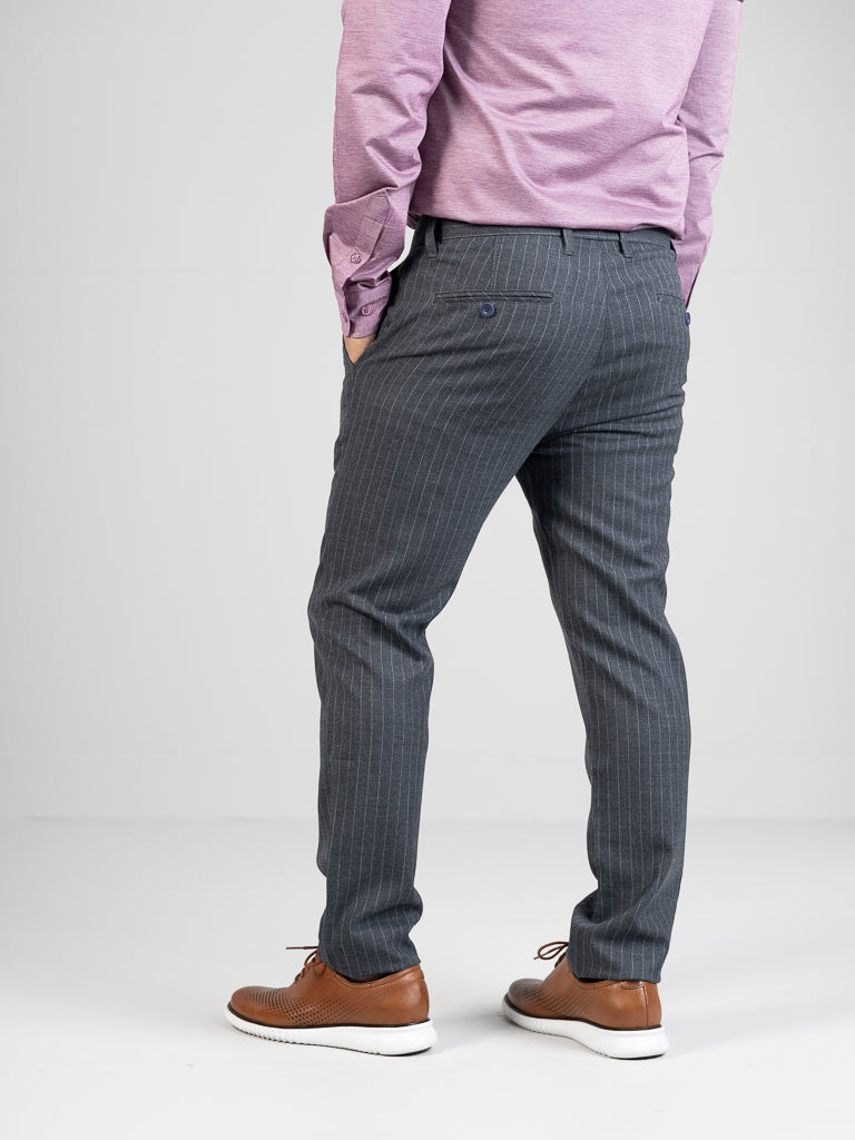 A person in a pink long-sleeve shirt and ID Menswears Hudson Wool Blend Pin Stripe Pant is facing away. These gray, mid-rise, slim-fit trousers are paired with brown shoes with white soles against a plain white background, with one hand casually in their pocket.