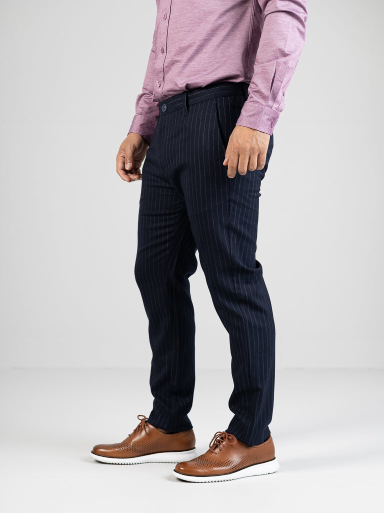 Against a plain white background, a person in a mauve long-sleeve shirt showcases ID Menswears Hudson Wool Blend Pin Stripe Pant in navy. The mid-rise slim-fit pants are paired with brown leather shoes featuring white soles, focusing on the lower half of the body.