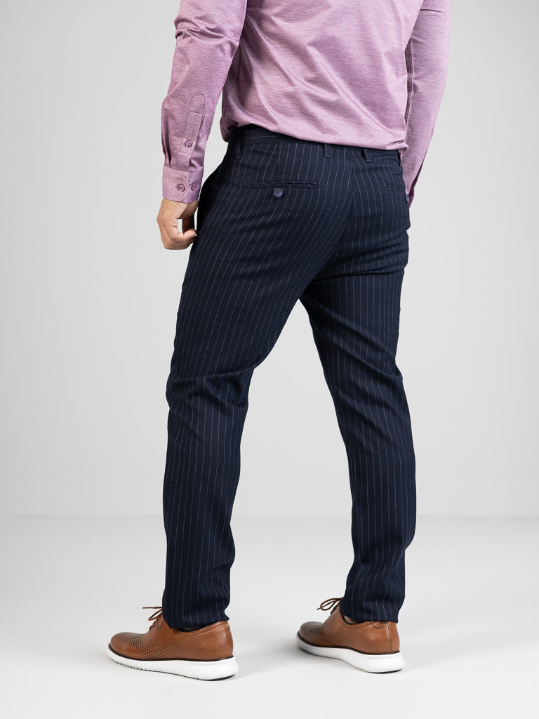 Seen from the back, a person wears a pink long-sleeve shirt and ID Menswears Hudson Wool Blend Pin Stripe Pants. Their hands rest at their sides. They wear brown dress shoes with white soles, exuding sophistication against the plain gray background.