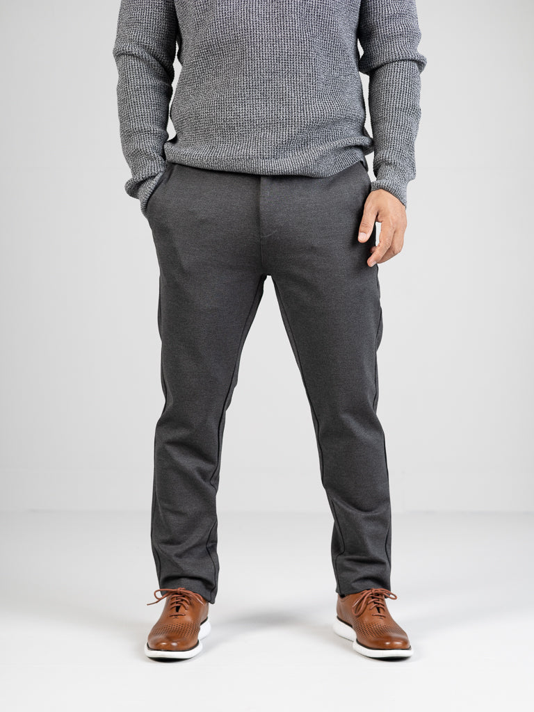 A person in a gray sweater and ID Menswears Manhattan 4-way Stretch Dress Pant stands hands in pockets, complemented by light brown shoes with white soles against a plain light gray background, creating a sleek business casual look.