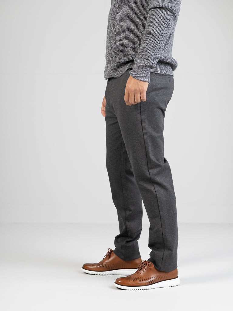 A person stands against a plain white background, wearing a wrinkle-resistant gray sweater, Manhattan 4-way Stretch Dress Pant by ID Menswear in slim-fit, and brown shoes with white soles. The focus is on the clothing, not the visible face.