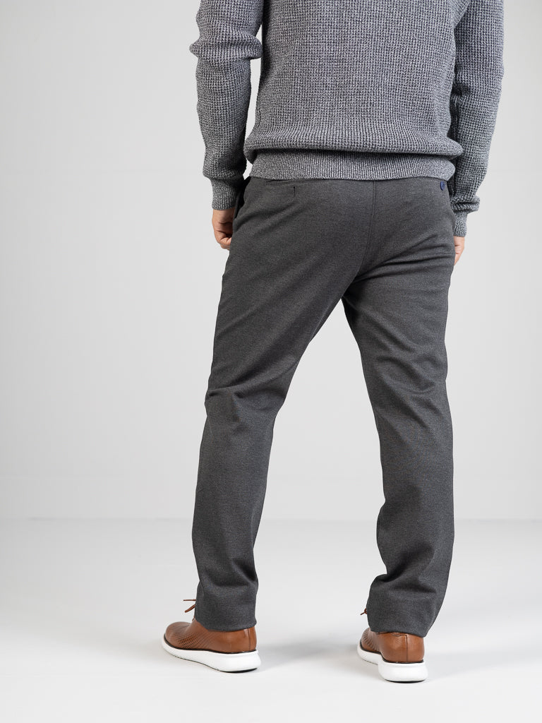 A person stands back to the camera in a gray sweater and ID Menswears Manhattan 4-way Stretch Dress Pant, exuding a business-casual vibe against a plain white wall, paired with brown sneakers.