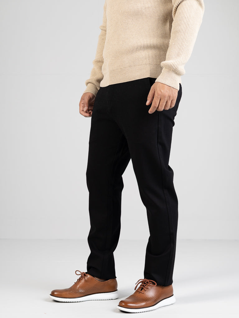 A person stands against a plain background, wearing a beige sweater, Manhattan 4-way Stretch Dress Pant in black from ID Menswear, and brown leather shoes with white soles. The image highlights the wrinkle-resistant outfit from shoulders down, perfectly capturing a business casual style.