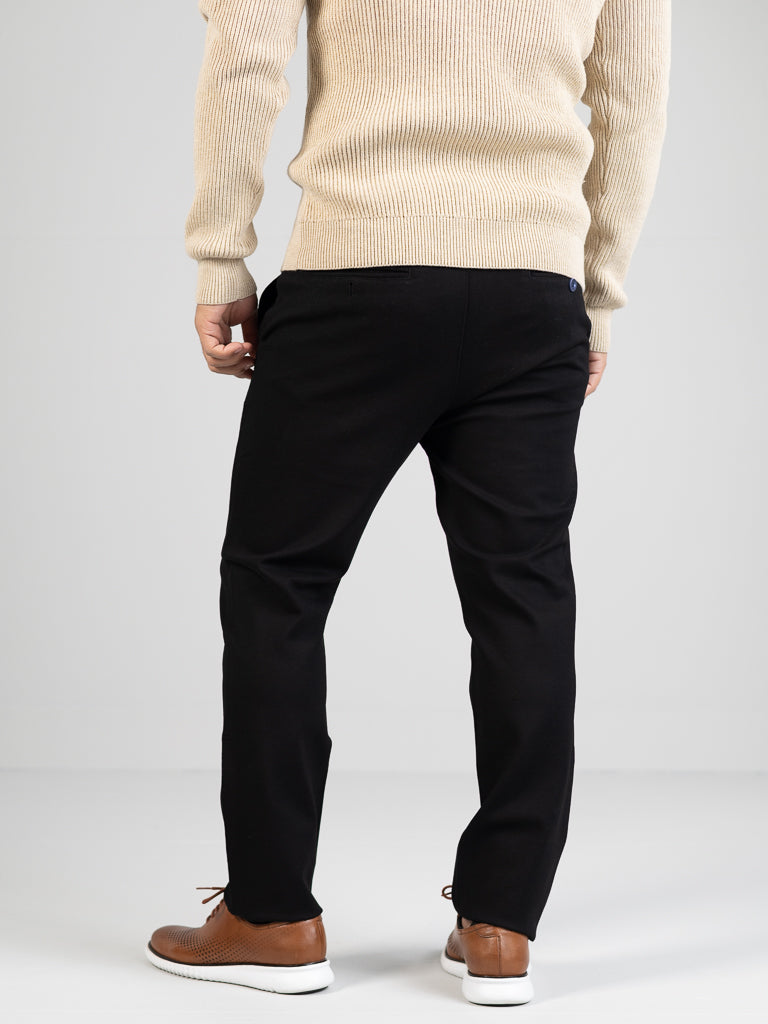 A person stands with their back to us, wearing a beige sweater and ID Menswears Manhattan 4-way Stretch Dress Pant in black, paired with brown dress shoes. The ensemble gives off a business casual vibe against the grayish-white backdrop.
