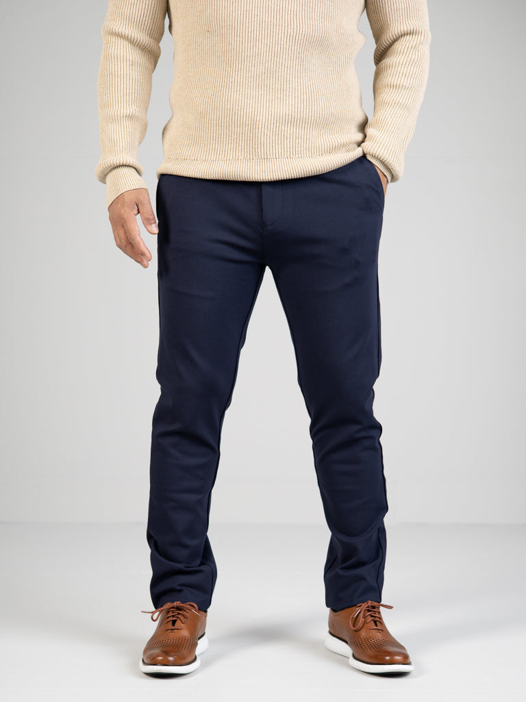 A person stands against a plain background wearing a beige sweater, ID Menswears Manhattan 4-way Stretch Dress Pant (slim-fit and wrinkle-resistant), and brown leather shoes. The focus is on the business casual outfit from neck to feet.