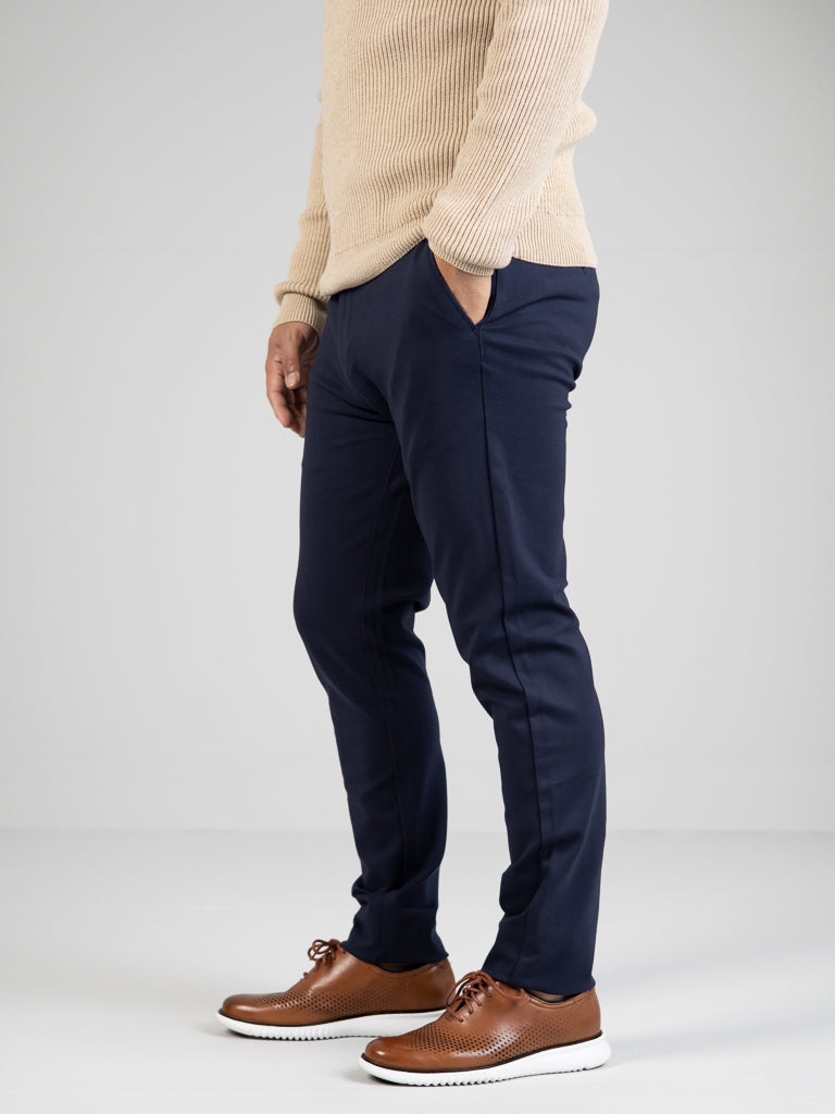 Dressed in a beige sweater, Manhattan 4-way Stretch Dress Pant by ID Menswear, and brown leather shoes with white soles, a person stands hands in pockets against a plain backdrop, perfectly blending style and wrinkle-resistant business casual elegance.