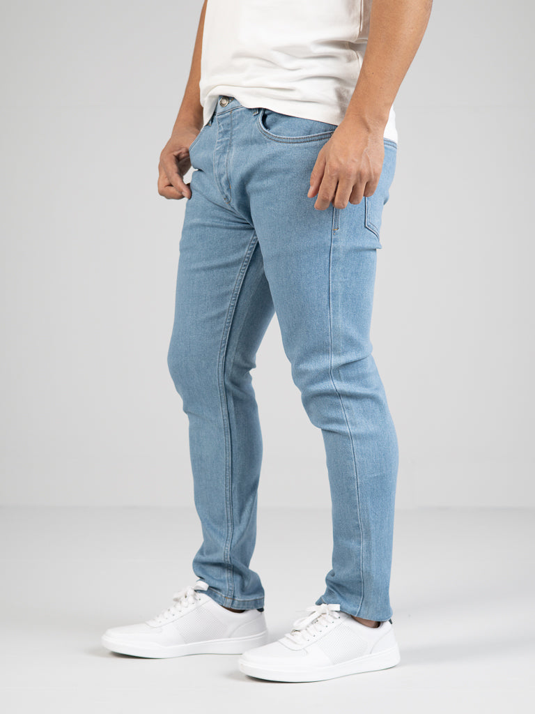 A person in Ganbaru Stretch Jean by ID Menswear stands against a gray background. They wear light blue denim jeans with a slim-tapered fit, paired with a white shirt and white sneakers, highlighting the cotton twill fabrics stylish cut and feel of the jeans.