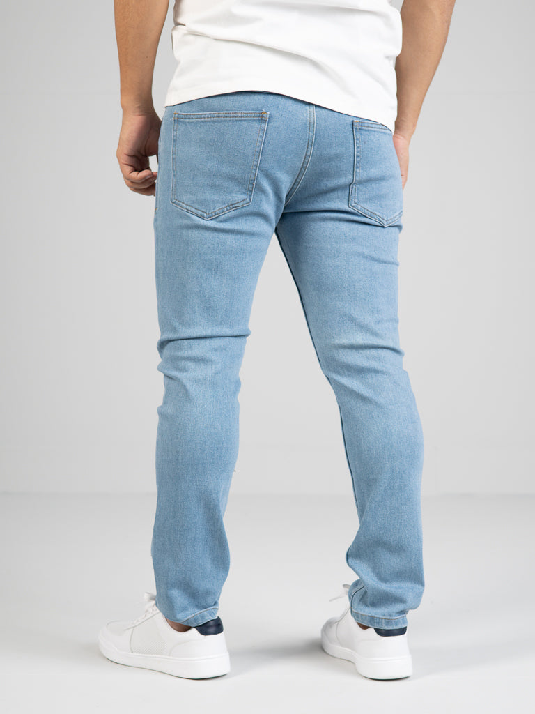 A person, wearing the light blue Ganbaru Stretch Jean by ID Menswear and white sneakers, stands on a white background facing away. These classic fit jeans, made from cotton twill, include back pockets that enhance their timeless appeal.
