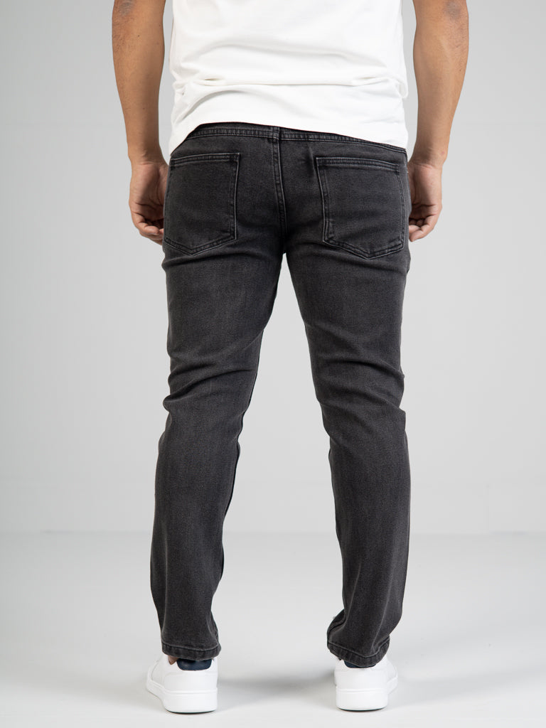 A person stands facing away in ID Menswears Ganbaru Stretch Jean, a slim-tapered fit in gray, paired with a white shirt and white sneakers. The plain light-colored backdrop highlights the sleek design of their cotton twill outfit.