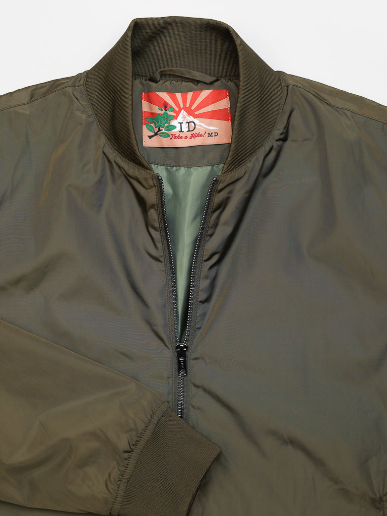 The olive green Catskill Bomber Jacket by ID Menswear has a zippered front, water-resistant shell, lighter green lining, and ribbed collar and cuffs. Inside, a red art deco tag with a rose reads Take a Hike! MD.