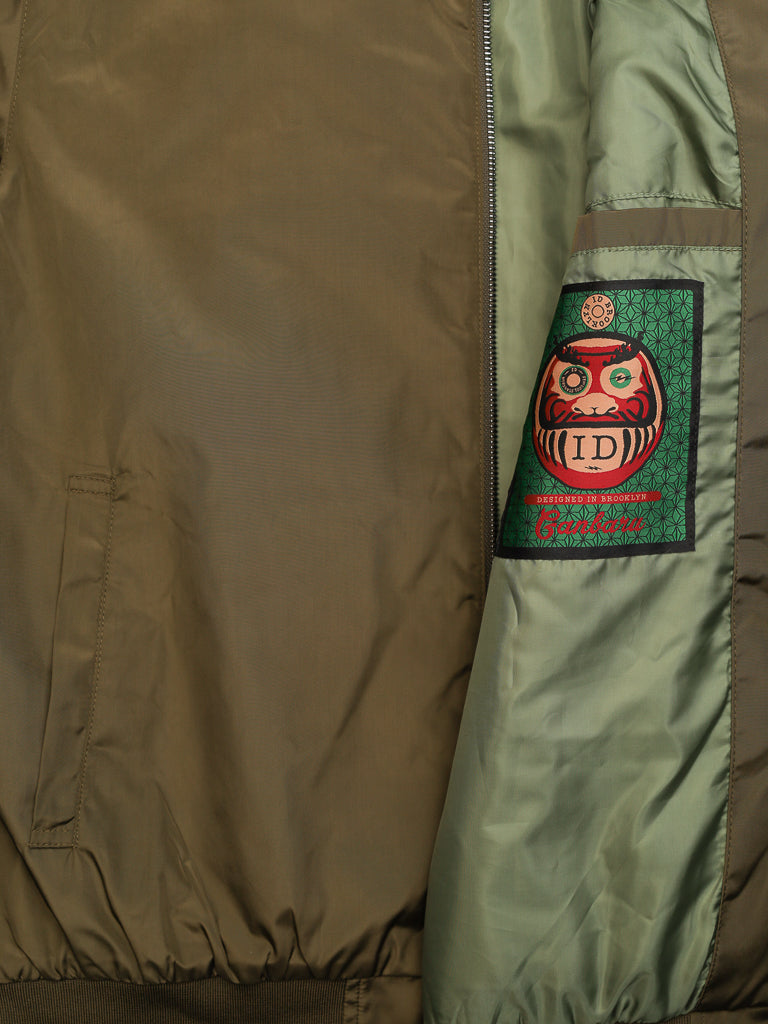 The olive green Catskill Bomber Jacket by ID Menswear features a partially visible sleeve patch with a red Daruma doll design and ID and Culture text. Its water-resistant lighter green inner lining adds practicality to its stylish appearance.