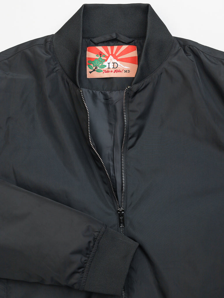 The Catskill Bomber Jacket by ID Menswear features a water-resistant black design with a front zipper, ribbed collar and cuffs. Inside, youll find a vibrant red and white sunburst pattern paired with a green and red artistic design, plus Take a Trip! MD text for added style and function.