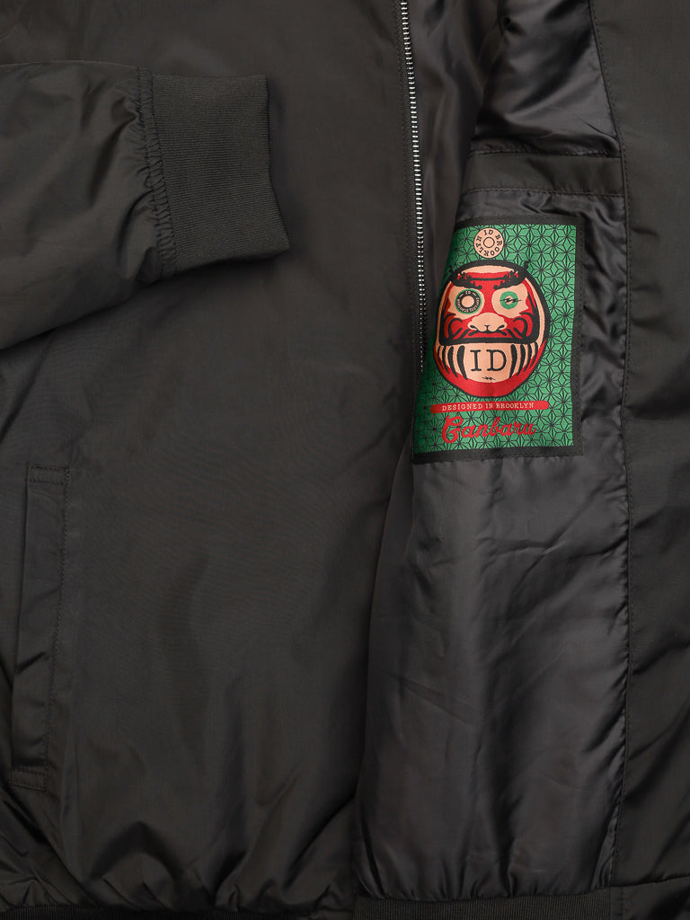 Partially unzipped Catskill Bomber Jacket by ID Menswear reveals its interior label, which features a vibrant design with a traditional Daruma doll against an intricate green background. This black, lightweight, water-resistant jacket also exposes one arm sleeve.