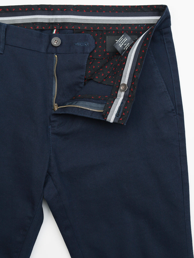 The Wythe Twill Stretch Chino by ID Menswear is a navy blue slim-fit pant featuring an open zipper and button that reveals a black and red arrow patterned waistband and pocket lining. Made from cotton twill, its ideal for casual occasions, with a clothing tag visible inside the waistband.