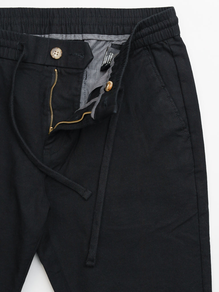 The Kent Drawstring Canvas Pant by ID Menswear is a slim-fit, mid-rise design in black. It features an elastic waistband with drawstring, gold zipper with tan button detailing, side pockets, and a visible gray inner lining at the waist, crafted from cotton canvas.