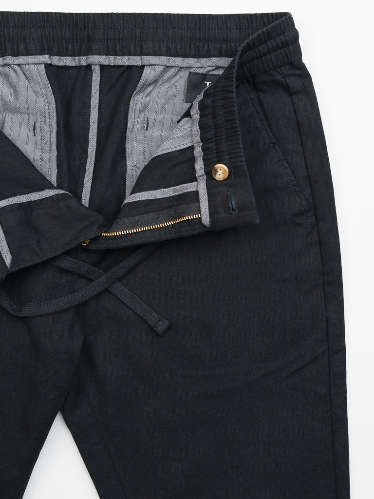 The Kent Drawstring Canvas Pant by ID Menswear is a slim-fit, mid-rise black pant featuring an open zipper and button fly. Made from cotton canvas, it has an elastic waistband with gray interior lining and a practical drawstring for comfort and style.