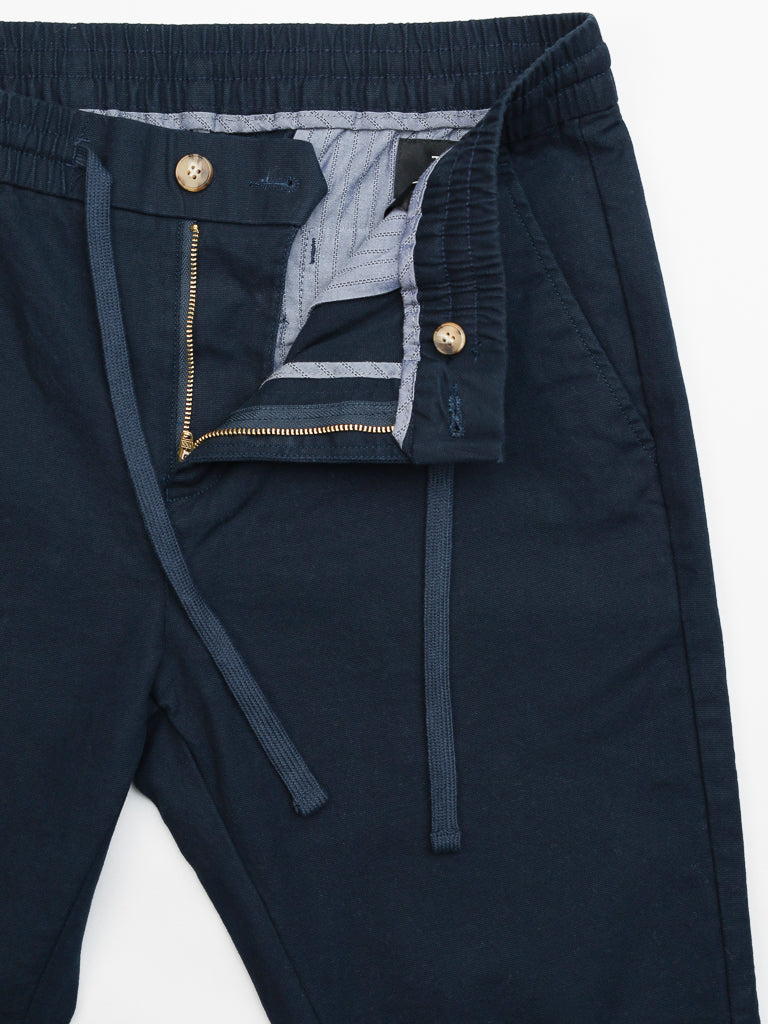 The Kent Drawstring Canvas Pant by ID Menswear in navy blue features a cotton canvas construction with an elastic waistband, slim-fit mid-rise cut, and button and zipper fly revealing interior lining. It includes two wooden buttons for added detail.