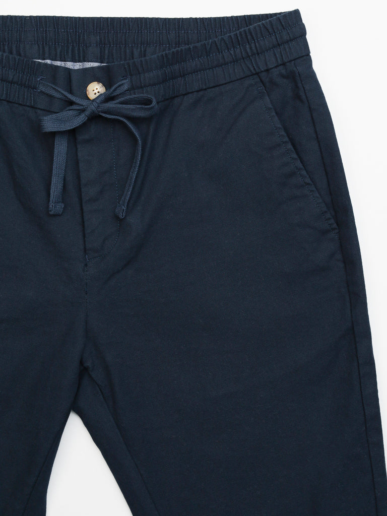 The Kent Drawstring Canvas Pant by ID Menswear is navy blue, slim-fit, and mid-rise. It features an elastic waistband with a drawstring and front pockets with a button detail, offering comfort and style.