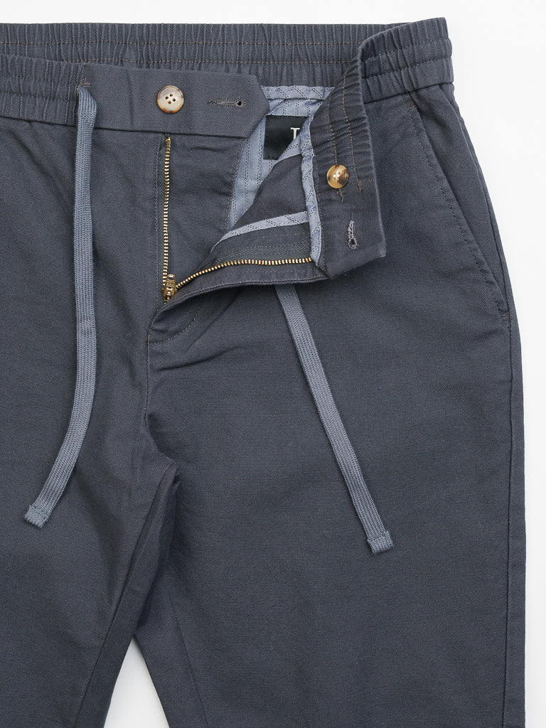 The Kent Drawstring Canvas Pant by ID Menswear features gray slim-fit mid-rise cotton canvas pants with an elastic waistband, button above a partially open zipper, front pockets, and visible hem.
