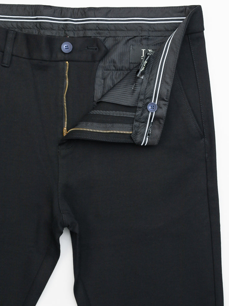 The Manhattan 4-way Stretch Dress Pant by ID Menswear is a black slim-fit style with a visible golden zipper and dark button closure. White and gray striped waistband features belt loops and side pockets. Its wrinkle-resistant fabric suits business casual settings, and the inside label displays a pattern.