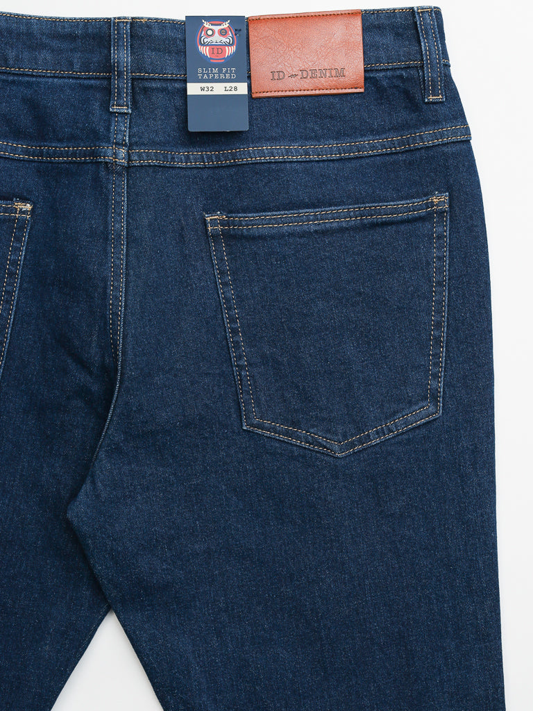 Close-up of Ganbaru Stretch Jean by ID Menswear: dark blue slim-tapered fit with visible stitching, back pocket, and brown leather waistband patch. A blue branded tag is near the pocket.
