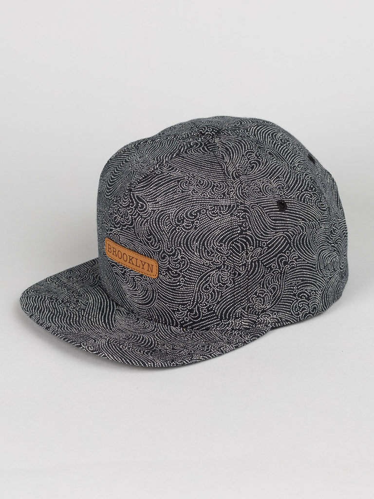 The Japanese Printed Nami Cotton Trucker cap by ID Menswear is gray, made of 100% cotton, with a swirling pattern and a tan leather patch labeled BROOKLYN. It features a flat brim and is displayed on a plain white background.