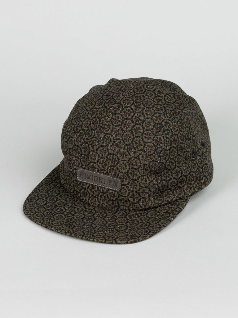 The Japanese Print Camp Hat by ID Menswear is a dark brown baseball cap with a geometric pattern, crafted from Japanese cotton. It features a flat brim and a Brooklyn leather patch on the front, made in Brooklyn against a plain white background.