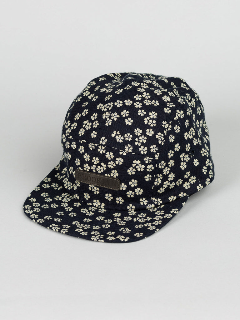 The Japanese Print Camp Hat by ID Menswear is a black 5-panel cap with a flat brim, crafted from Japanese printed cotton featuring a small white floral pattern. It has a brown leather Made in Brooklyn patch on the front and is set against a plain white background, exuding modern charm.