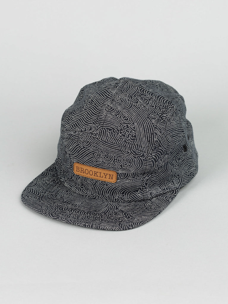 The Japanese Print Camp Hat by ID Menswear is a black and white patterned camp cap made from Japanese printed cotton, featuring a brown BROOKLYN leather patch on the front. Its proudly made in Brooklyn and displayed on a plain gray background.