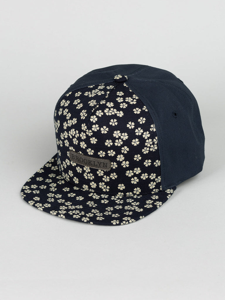 The ID Menswear Japanese Printed Sakura Cotton Trucker is a navy snapback cap made from 100% cotton, showcasing a beige floral pattern on the front panel and visor. It features a centered brown rectangular BROOKLYN patch embossed with flair on a light gray background.