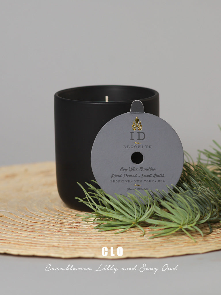 A black soy wax candle with a round, gray ID Menswear label sits on a woven mat, surrounded by pine branches. Text at the bottom reads CLO - Casablanca Lilly and Sexy Oud. This candle is handmade in Brooklyn, New York, USA.