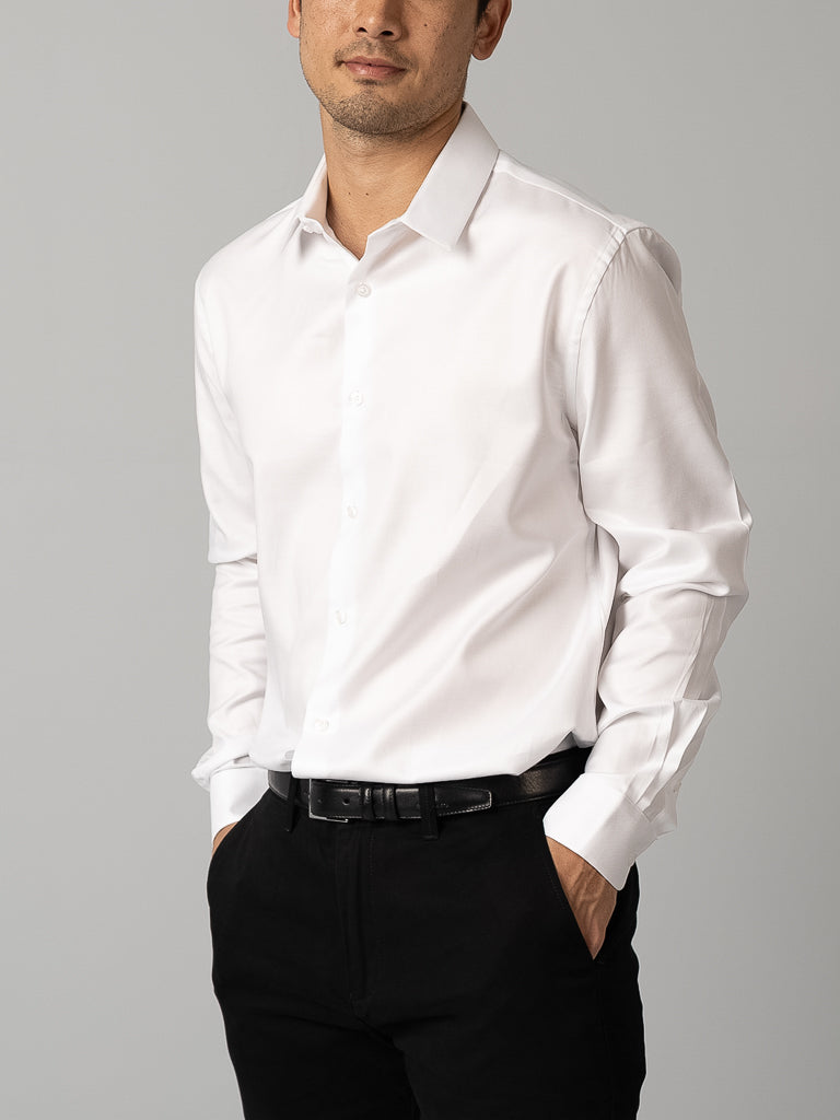 A man wears the ID Menswear Cotton Dress Shirt in slim fit, white, paired with black pants. He stands with hands in pockets against a gray background, his face partially visible and expression relaxed.