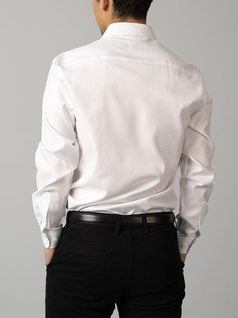 A man faces away from the camera, modeling the ID Menswear Cotton Dress Shirt in a slim fit—white, long-sleeved—paired with black pants and a belt, set against a plain light gray background.